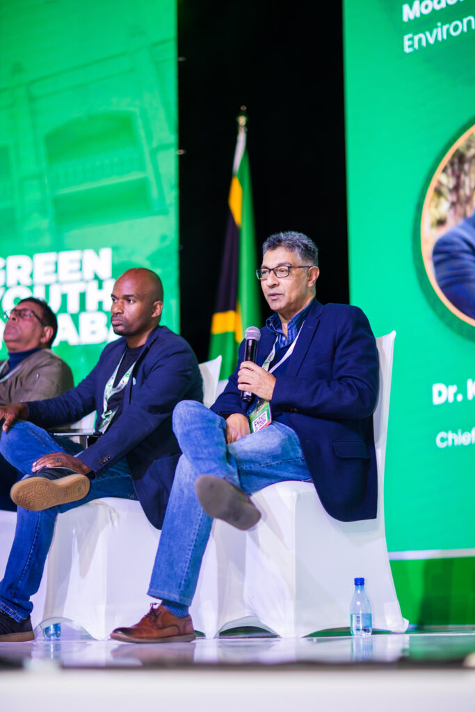 Panel of speakers seated on stage with green backdrop and large logos; man in blazer holding a microphone speaks while others listen.